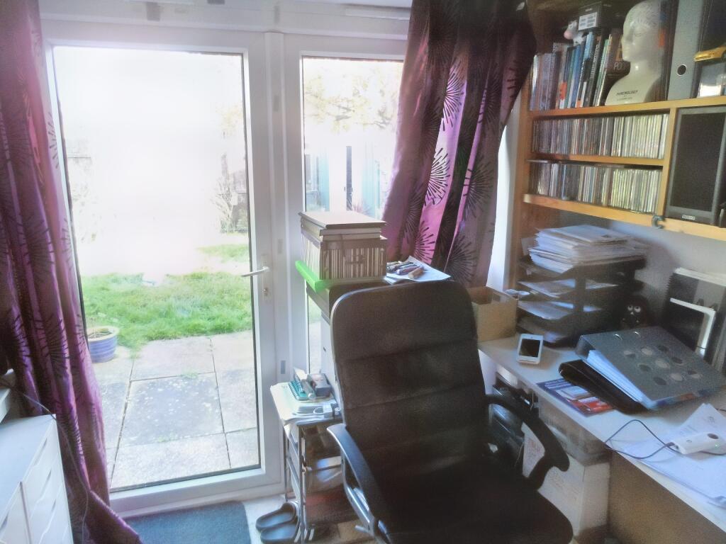 A compact home office at 1 Boscombe Close Lincoln features an ergonomic black chair facing a desk cluttered with documents, books, and electronic devices. Shelves with books and CDs are mounted on the wall. Large glass doors draped with purple curtains lead to a patio and garden outside. Now on the market with Palace Auctions.