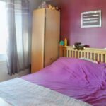 A bedroom at 1 Boscombe Close Lincoln features a wooden bed frame and a purple bedspread. A small stuffed animal rests on the bed. Next to it, a wooden wardrobe holds various stuffed animals on top. A framed picture adorns the purple wall, with light filtering through a partially open window. On the market with Palace Auctions.