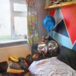 A bright and colorful bedroom at 1 Boscombe Close, Lincoln is on the market with Palace Auctions. It features a bed in the corner adorned with white bedding. Above the bed are geometric patterned walls. Balloons, one blue and the other silver, float nearby. Patterned curtains let in natural light while clothing is piled on the bed.