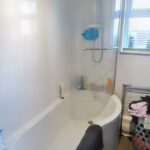 A modern bathroom at 1 Boscombe Close, Lincoln, boasting a curved shower-bathtub combination. The pristine white tub with a clear shower curtain includes toiletries neatly arranged on a small shelf. A towel drapes over the edge, while a laundry basket sits to the right beneath a window with blinds. Now on the market with Palace Auctions.