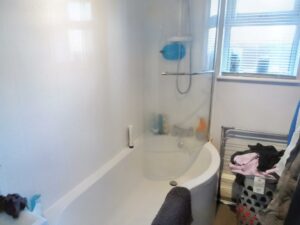 A modern bathroom at 1 Boscombe Close, Lincoln, boasting a curved shower-bathtub combination. The pristine white tub with a clear shower curtain includes toiletries neatly arranged on a small shelf. A towel drapes over the edge, while a laundry basket sits to the right beneath a window with blinds. Now on the market with Palace Auctions.