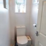 A small bathroom at 1 Boscombe Close Lincoln features a white toilet with a roll of toilet paper mounted on the wall to the right. Above the toilet, sunlight streams through a window with a potted plant on the sill. The door is partially open, highlighting its charm now on the market with Palace Auctions.