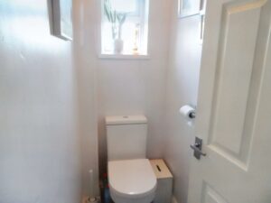 A small bathroom at 1 Boscombe Close Lincoln features a white toilet with a roll of toilet paper mounted on the wall to the right. Above the toilet, sunlight streams through a window with a potted plant on the sill. The door is partially open, highlighting its charm now on the market with Palace Auctions.