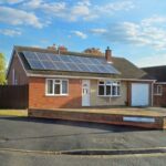 A single-story brick house with solar panels on the roof, a white front door, and an attached garage. Situated on the corner of Boscombe Close, Lincoln, this charming home is now on the market with Palace Auctions. A street lamp stands nearby under a clear, partly cloudy sky.