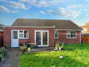 1 Boscombe Close Lincoln is on the market with Palace Auctions. This charming single-story brick house features a moss-covered, gabled roof, three white-framed windows, and a white patio door. The backyard boasts a small table with two chairs, potted plants, and a green lawn under a blue sky with scattered clouds.