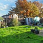 A backyard at 1 Boscombe Close, Lincoln, featuring a lush green lawn, several potted plants, and a cozy patio area. A large, leafy tree with autumn foliage stands tall in the background. There's also a shed and greenhouse visible, with neighboring houses and a clear sky enhancing the serene setting. Now on the market with Palace Auctions.