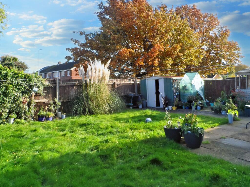 A backyard at 1 Boscombe Close, Lincoln, featuring a lush green lawn, several potted plants, and a cozy patio area. A large, leafy tree with autumn foliage stands tall in the background. There's also a shed and greenhouse visible, with neighboring houses and a clear sky enhancing the serene setting. Now on the market with Palace Auctions.