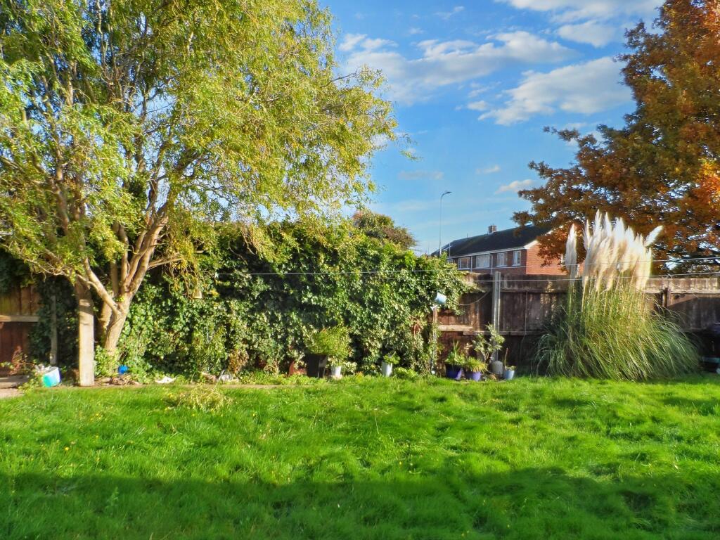 A backyard at 1 Boscombe Close, Lincoln, features a lush green lawn bordered by various plants and trees. A large tree on the left has ivy covering parts of the fence behind it. To the right, there is a tall ornamental grass plant. Some houses are visible in the background under a partly cloudy sky.