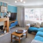 A living room at 1 Boscombe Close, Lincoln, features a beige sofa with blue throws, a wooden coffee table, and a TV mounted above a blue and wood TV unit. The fireplace boasts a wooden mantel adorned with art pieces and plants. A large window with white curtains lets in natural light—a must-see on the market with Palace Auctions.