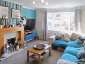 A living room at 1 Boscombe Close, Lincoln, features a beige sofa with blue throws, a wooden coffee table, and a TV mounted above a blue and wood TV unit. The fireplace boasts a wooden mantel adorned with art pieces and plants. A large window with white curtains lets in natural light—a must-see on the market with Palace Auctions.