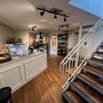 A cozy coffee shop at 120 St Margaret's Road, Twickenham, features an inviting interior with a counter displaying pastries, a coffee machine, and shelves with products. Wooden floors and white walls complement the staircase with white railings leading to an upper level. Track lighting creates a warm ambiance.