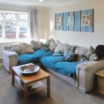 A cozy living room at 1 Boscombe Close, Lincoln, featuring a large L-shaped beige sofa with blue throws and numerous cushions. A wooden coffee table with a decorative bowl stands in front of the sofa. The room is brightened by a window with light curtains, and a plant decorates a side table. Now on the market with Palace Auctions!
