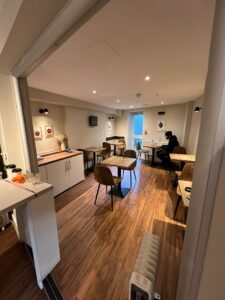 A small, cozy cafe at 120 St Margret's road Twickenham, with wooden flooring and several tables and chairs arranged neatly. Two people are sitting at separate tables engrossed in their activities. The walls are decorated with simple art pieces, and there's a small counter with items on it to the left.