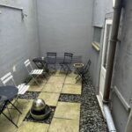 A small, enclosed outdoor patio space at 120 St Margret's Road, Twickenham, with a gray concrete floor and walls. It features metal patio furniture, including two round black tables and several white and black chairs. A spherical decorative item takes center stage, surrounded by paving stones and gravel.