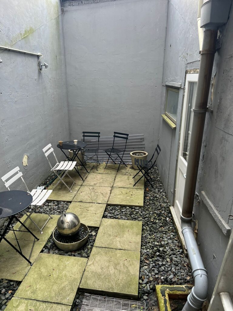 A small, enclosed outdoor patio space at 120 St Margret's Road, Twickenham, with a gray concrete floor and walls. It features metal patio furniture, including two round black tables and several white and black chairs. A spherical decorative item takes center stage, surrounded by paving stones and gravel.