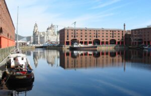 Property for sale by Auction The image shows a scenic view of Albert Dock in Liverpool, with historic red-brick warehouses reflected in the calm water. Several boats are moored along the dock, and the skyline features iconic buildings under a clear blue sky. Presented by Palace Auctions