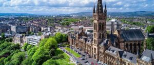 Aerial view of the cityscape with historical buildings and modern structures. Prominently featured is a large Gothic architecture building with a tall clock tower, set amidst lush green trees. For those interested, Glasgow Property Investment Guide highlights this area's potential amidst sprawling urban landscapes and distant hills.