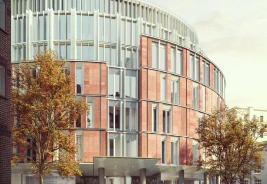 The future Harrods conneted hotel Modern building with a curved facade, featuring red brickwork and vertical white elements, surrounded by autumnal trees