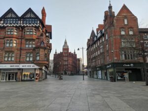 A wide, empty street is flanked by historic, red-brick buildings with ornate architectural details. Shops, including "White Rose" and "Clarks," have signs along the ground floors. In the distance, a large, castle-like structure is centered at the end of the street.