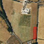 Aerial view of farmland with a marked red rectangular plot on the right side. The plot is adjacent to a road labeled “Beech Farm Rd” and is surrounded by larger empty fields. Several buildings and structures are visible at the top center, with trees lining some field borders.