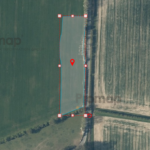 Aerial view of agricultural fields with a marked polygonal area highlighted in blue, outlined in red, featuring a red pin in the center. The highlighted area spans a section of the field and is bordered by a line of trees on the right-hand side.