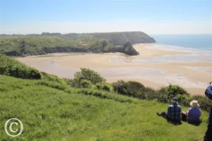 A scenic coastal view features a wide sandy beach framed by lush green cliffs under a clear blue sky. In the foreground, two people sit on the grassy hill, enjoying the panoramic vista at Nicholaston House wedding venue.