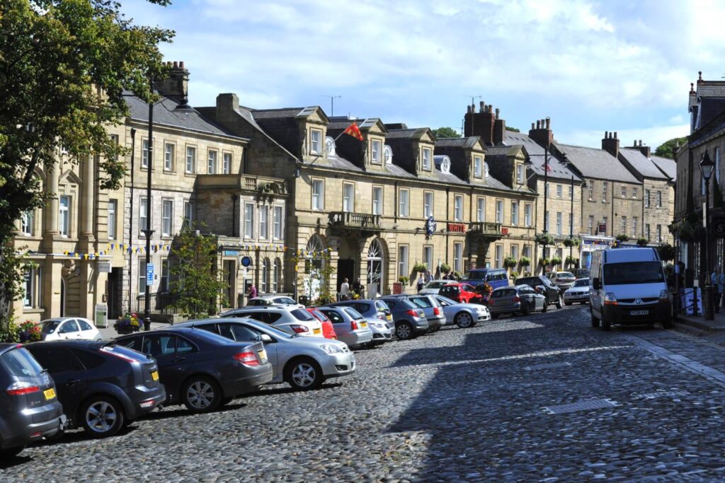 A cobblestone street lined with parked cars is flanked by historic stone buildings adorned with flags and hanging plants. A tree on the left provides shade, while a clear blue sky overhead suggests a pleasant day. Sidewalks are visible, adding to the quaint town atmosphere.