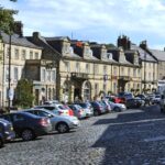 A cobblestone street lined with parked cars is flanked by historic stone buildings adorned with flags and hanging plants. A tree on the left provides shade, while a clear blue sky overhead suggests a pleasant day. Sidewalks are visible, adding to the quaint town atmosphere.