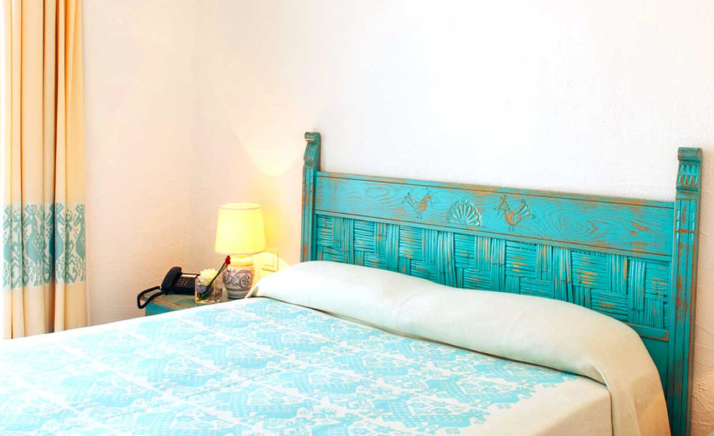 A cozy bedroom features a bed with a turquoise headboard and a matching bedspread adorned with intricate patterns. A beige blanket is folded at the foot of the bed. Beside the bed is a small nightstand with a lamp, a phone, and some decorative items.