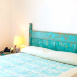 A cozy bedroom features a bed with a turquoise headboard and a matching bedspread adorned with intricate patterns. A beige blanket is folded at the foot of the bed. Beside the bed is a small nightstand with a lamp, a phone, and some decorative items.
