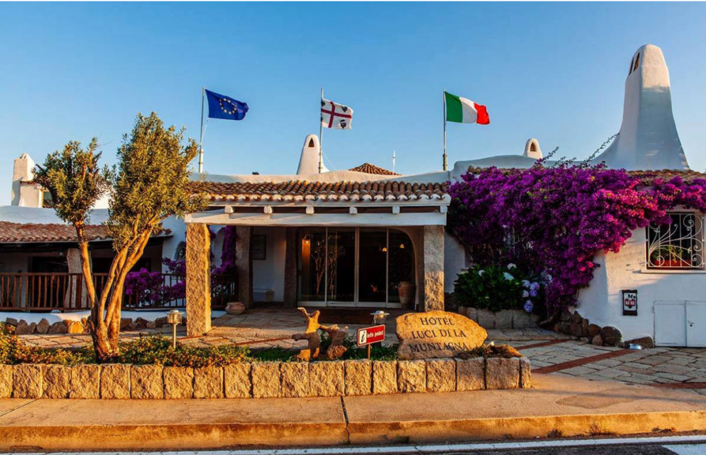 A charming hotel entrance with a stone pathway, colorful bougainvillea climbing the walls, and flags from different countries displayed on the roof. A welcoming stone sign reads "Hotel Lucci Della Montagna." The scene is bathed in warm sunlight.