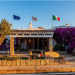 A charming hotel entrance with a stone pathway, colorful bougainvillea climbing the walls, and flags from different countries displayed on the roof. A welcoming stone sign reads "Hotel Lucci Della Montagna." The scene is bathed in warm sunlight.