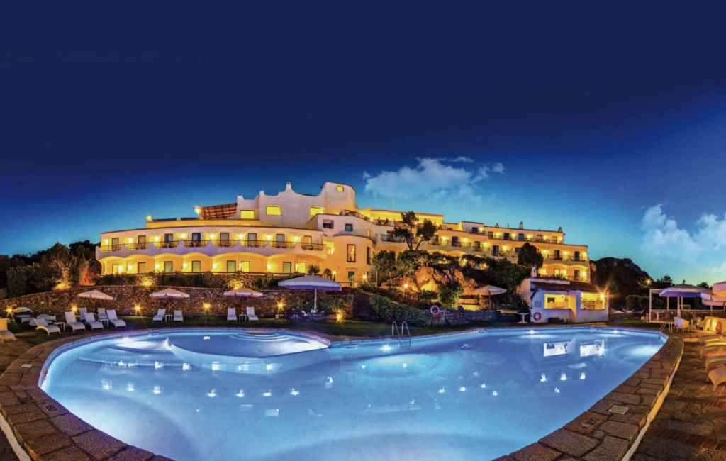 A luxurious hotel with bright lights illuminates the night sky, reflecting off a large, curvy swimming pool in the foreground. The hotel features multiple balconies and terraces. Sun loungers and umbrellas are arranged neatly around the pool area.