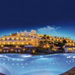 A luxurious hotel with bright lights illuminates the night sky, reflecting off a large, curvy swimming pool in the foreground. The hotel features multiple balconies and terraces. Sun loungers and umbrellas are arranged neatly around the pool area.