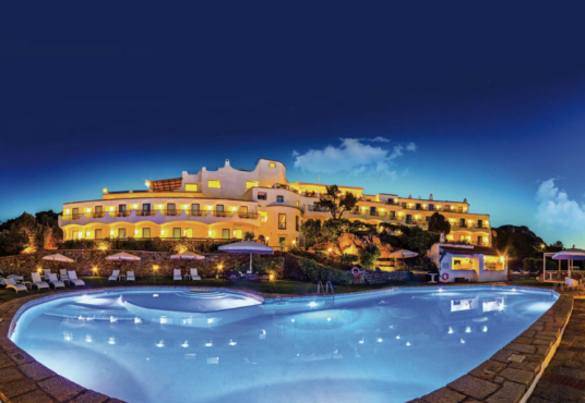 A luxurious hotel with bright lights illuminates the night sky, reflecting off a large, curvy swimming pool in the foreground. The hotel features multiple balconies and terraces. Sun loungers and umbrellas are arranged neatly around the pool area.