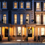 A street view at dusk shows elegant, illuminated townhouses with large windows and classic architectural details. Topiary plants in pots decorate the entrance, and a figure walks by on the sidewalk. Cars are parked along the street.