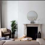 A modern living room featuring a round mirror above a decorative fireplace, a potted plant, and an armchair with a cushion. A beige sectional sofa faces a wooden coffee table with a magazine. The space includes neutral tones, wood flooring, and large windows with grey curtains.