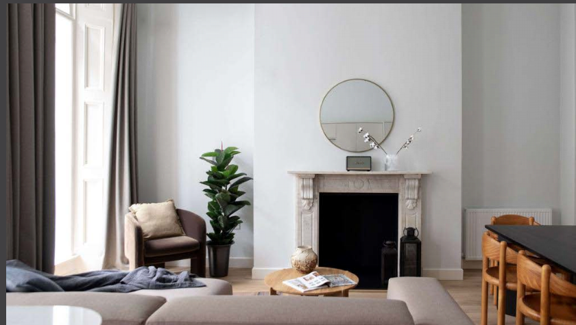 A modern living room featuring a round mirror above a decorative fireplace, a potted plant, and an armchair with a cushion. A beige sectional sofa faces a wooden coffee table with a magazine. The space includes neutral tones, wood flooring, and large windows with grey curtains.