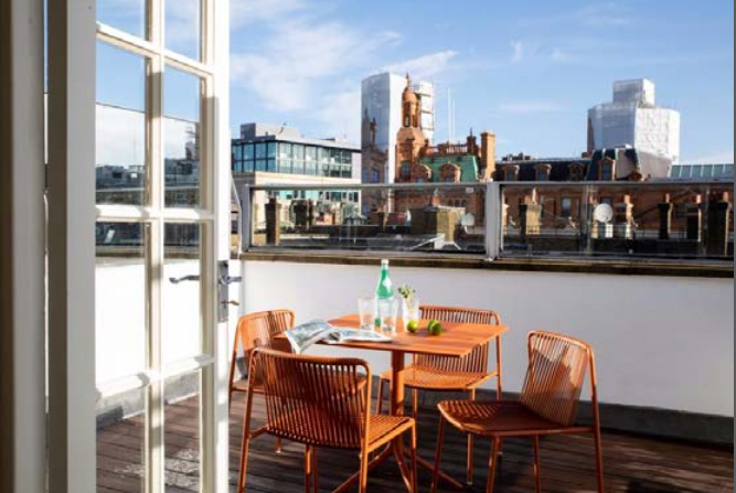 A sunny rooftop terrace features an orange outdoor dining set with four chairs and a round table. The table holds a bottle of water, a glass, and a few other small items. The terrace overlooks a cityscape with various buildings under a clear blue sky.
