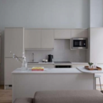 A modern kitchen with minimalist design features white cabinetry, an island with a sink, a built-in microwave, and a stove. A wooden table with chairs is set near the island. Light gray walls and neutral-colored decor create a clean, open space. There are stairs to the left.