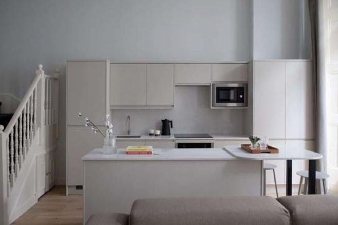 A modern kitchen with minimalist design features white cabinetry, an island with a sink, a built-in microwave, and a stove. A wooden table with chairs is set near the island. Light gray walls and neutral-colored decor create a clean, open space. There are stairs to the left.