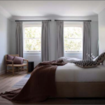A neatly arranged bedroom features a large bed with white linens and a brown throw. Two windows with grey curtains allow natural light to stream in. A wooden chair with cushions is in the corner, and a piece of artwork hangs on the wall. The room has a serene ambiance.