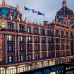 A large, ornate building is illuminated with numerous lights and grand architectural details. The facade features the name "Harrods" in bright lights. Flags are mounted on the roof, and the scene appears to be taken during twilight.