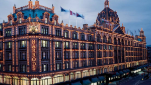 A large, ornate building is illuminated with numerous lights and grand architectural details. The facade features the name "Harrods" in bright lights. Flags are mounted on the roof, and the scene appears to be taken during twilight.