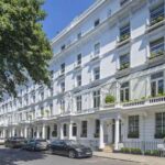 A row of elegant white terraced houses with tall windows and ornate balconies on a sunny day. Some houses have potted plants on their balconies. Several cars are parked along the street, and a large tree provides shade on the left—truly a Knightbridge development opportunity by Remi Charles.