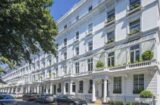 A row of elegant white terraced houses with tall windows and ornate balconies on a sunny day. Some houses have potted plants on their balconies. Several cars are parked along the street, and a large tree provides shade on the left—truly a Knightbridge development opportunity by Remi Charles.