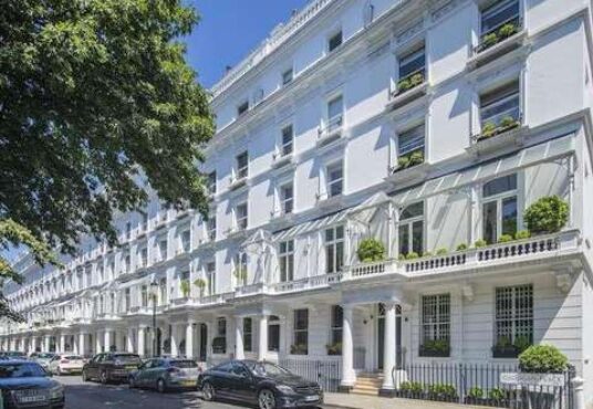 A row of elegant white terraced houses with tall windows and ornate balconies on a sunny day. Some houses have potted plants on their balconies. Several cars are parked along the street, and a large tree provides shade on the left—truly a Knightbridge development opportunity by Remi Charles.
