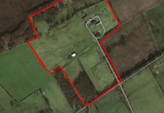 Aerial view of a large rural property outlined in red. The property includes several buildings and open, grassy fields bordered by trees and farmland. Surrounding areas are divided into different green fields with visible roads and paths.