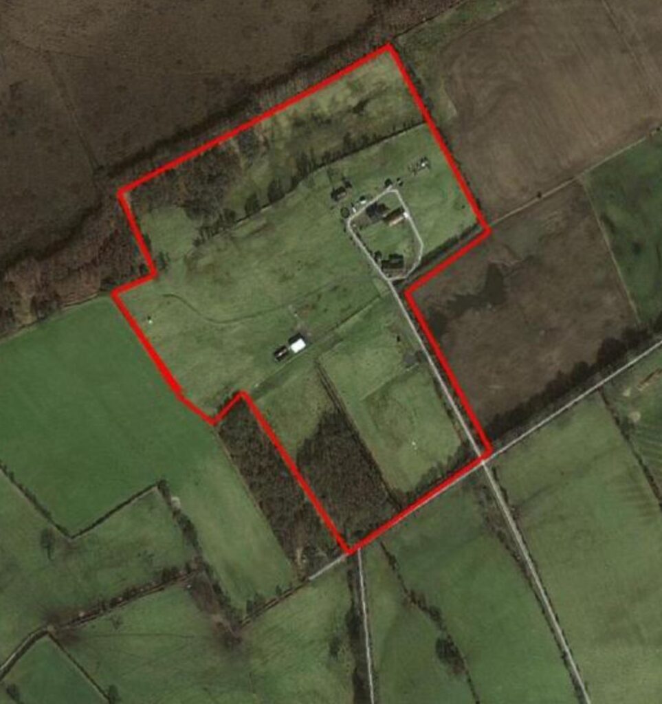 Aerial view of a large rural property outlined in red. The property includes several buildings and open, grassy fields bordered by trees and farmland. Surrounding areas are divided into different green fields with visible roads and paths.