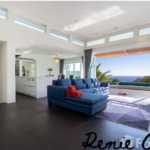 A modern, open-concept living room with a blue sectional sofa facing a large sliding glass door that opens to a scenic ocean view. The room has white walls, high windows, and a sleek kitchen area in the background. A chandelier hangs in the dining area.